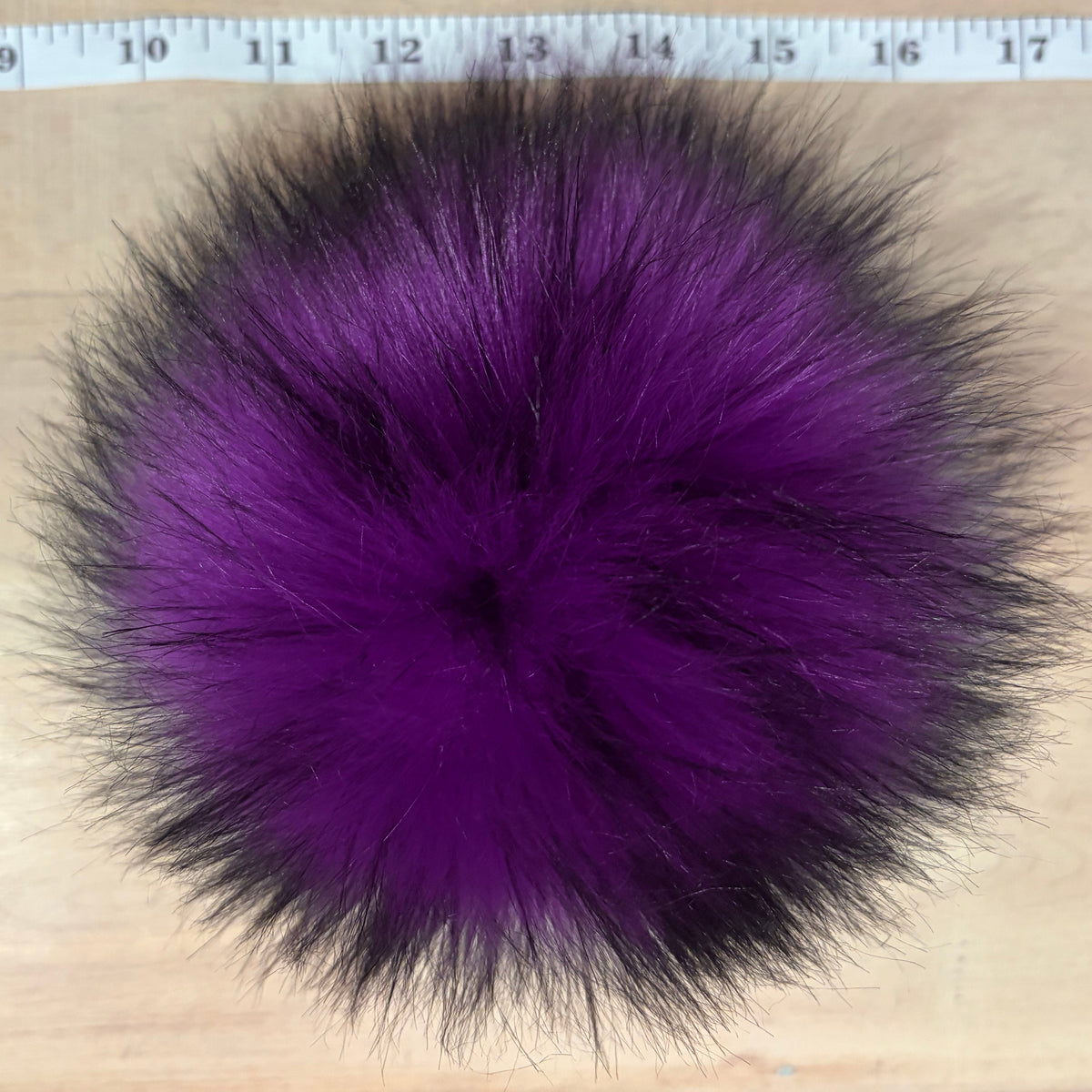 A fluffy pom-pom accessory in vibrant purple with black tips, shown on a light wood surface with a measuring tape visible at the top for scale.