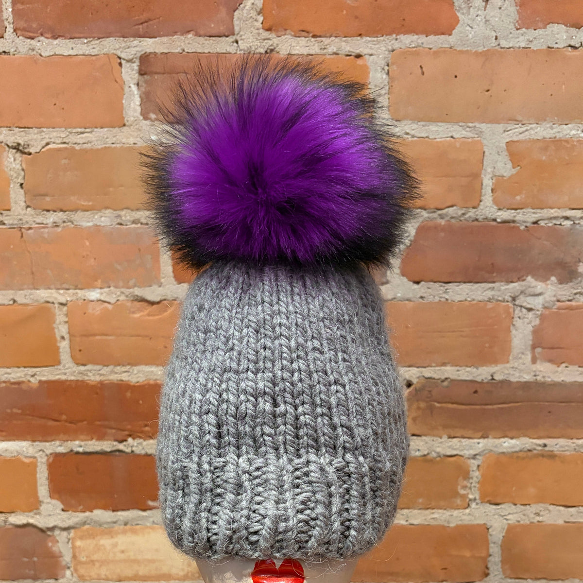 A gray knit beanie with a large fluffy purple pom-pom, displayed on a mannequin head against a brick wall background.