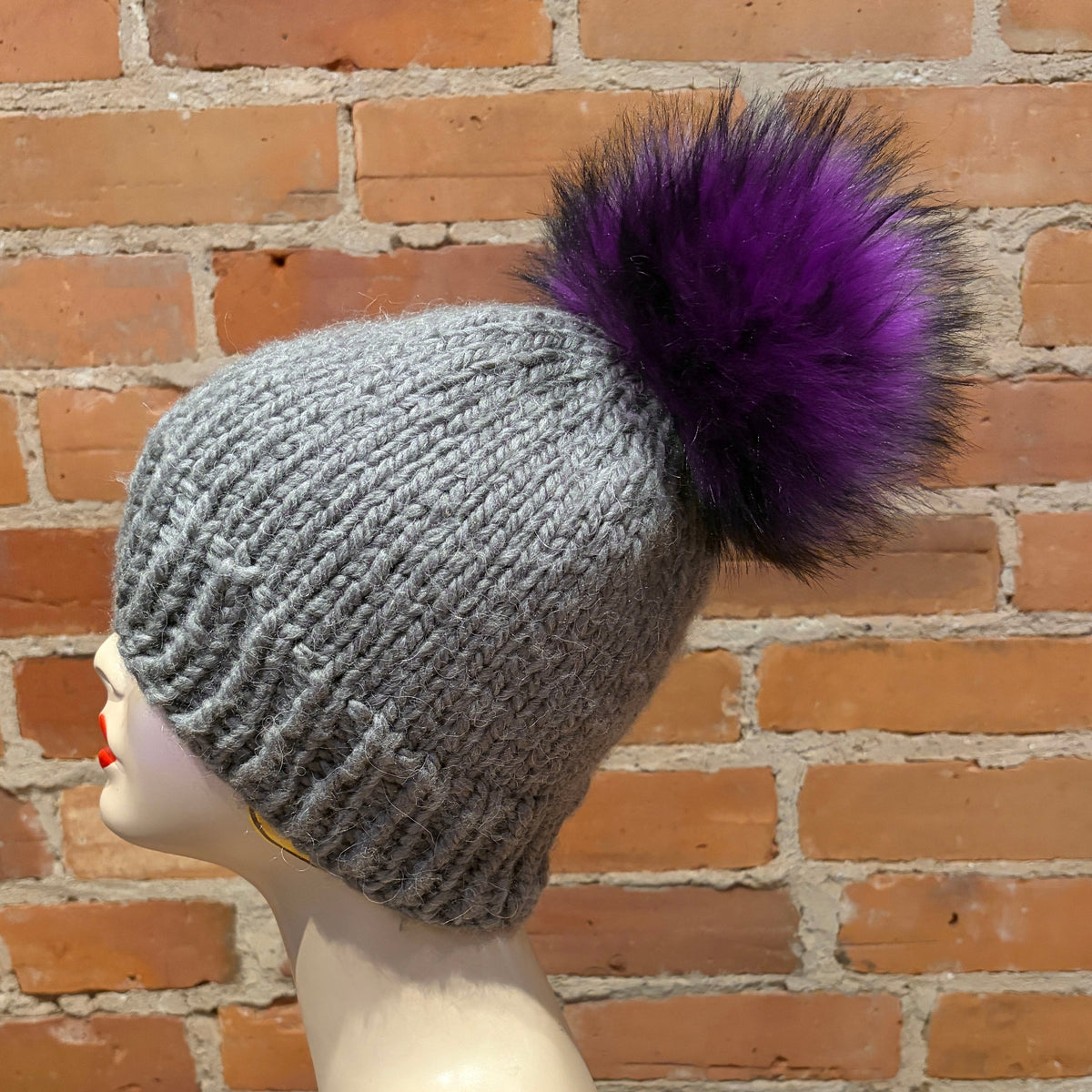 A gray ribbed knit beanie with a large fluffy purple pom-pom, displayed on a mannequin head against a brick wall background.