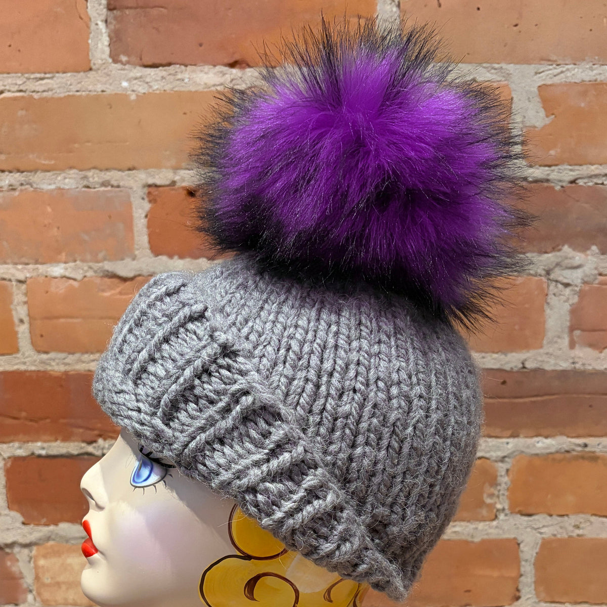 A gray knit beanie with a large fluffy purple pom-pom, displayed on a mannequin head against a red brick wall background.