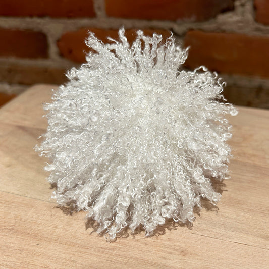 A fluffy, curly white decorative pom-pom or sculptural ornament made from textured synthetic fibers, displayed on a light wood surface with a blurred brick wall background.