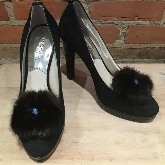A pair of black suede high-heeled pumps with fluffy black fur pom-poms and a small blue rhinestone accent, displayed on a light wood surface against a brick wall background.