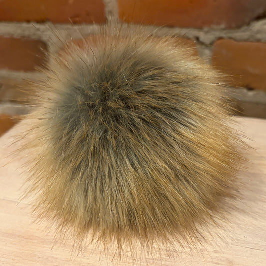 A round, fluffy faux fur pom-pom in a natural brown and beige color, resting on a light wood surface with a brick wall background.