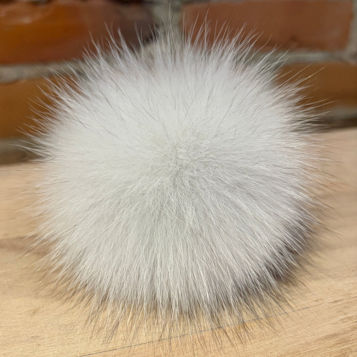 A fluffy spherical white fox fur pom-pom accessory resting on a light wood surface with a brick wall background.