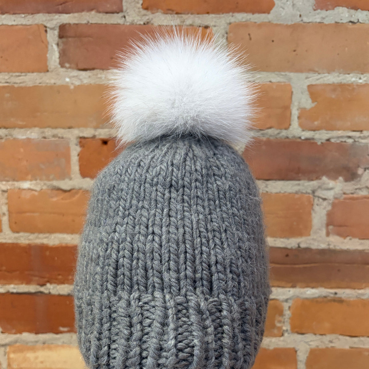 A gray ribbed knit beanie with a fluffy white fox-fur pom-pom, displayed on a mannequin head with red lips in front of a brick wall background.