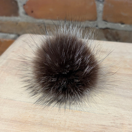 A round, fluffy raccoon-fur pom-pom in dark brown with lighter brown and gray tips, resting on a light wood surface with a brick wall in the background.