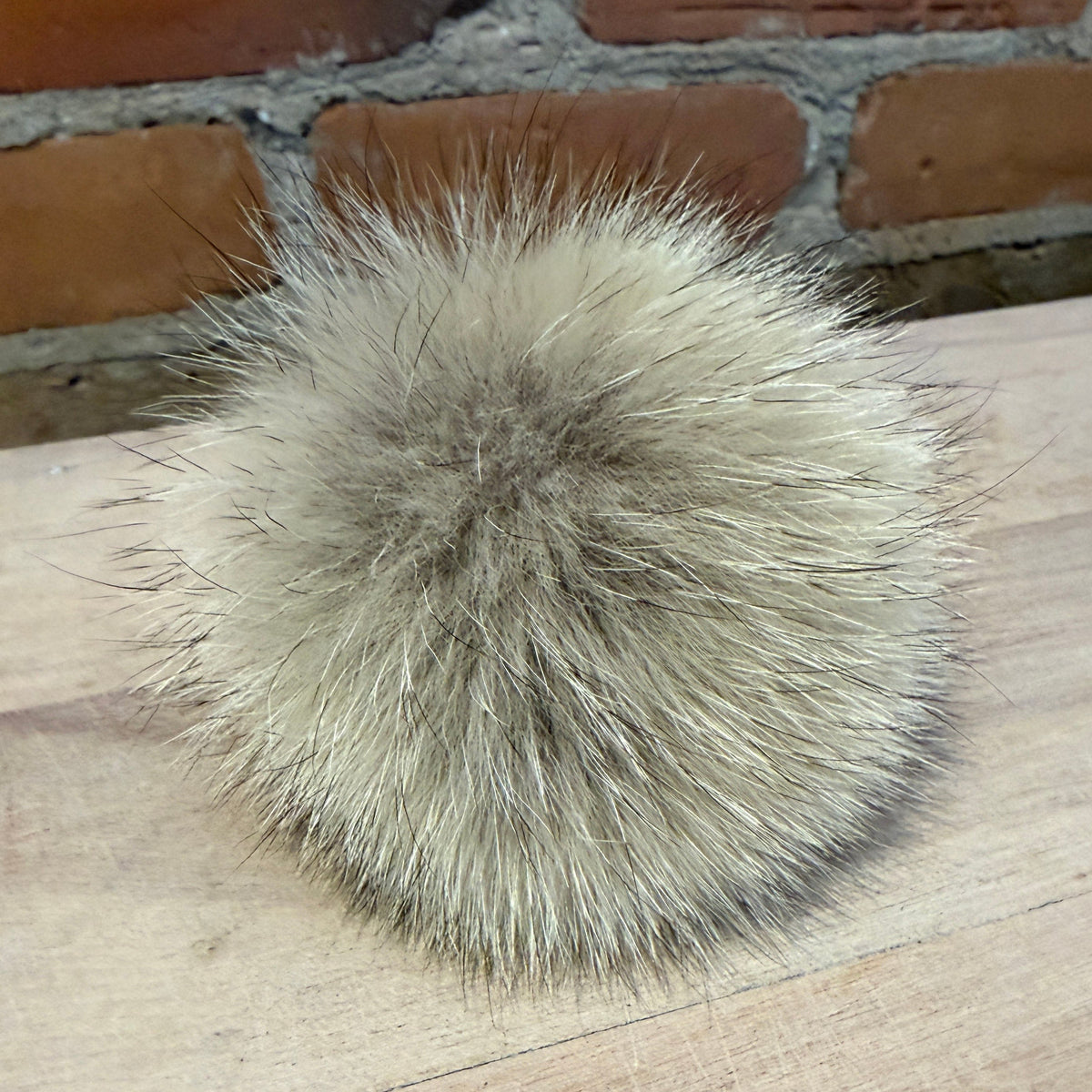A fluffy pom-pom accessory made of natural light brown and beige fur, resting on a light wood surface with a brick wall in the background.