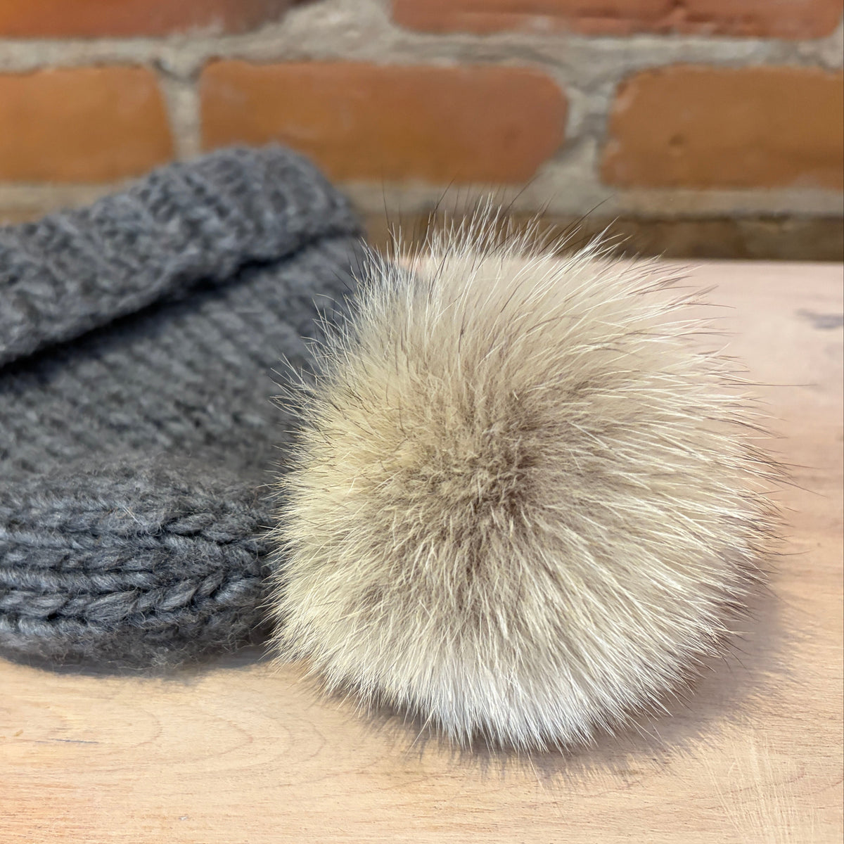 A small dark gray knit beanie hat with a small fluffy beige coyote-fur pom-pom, displayed on a light wood surface with a brick wall background.