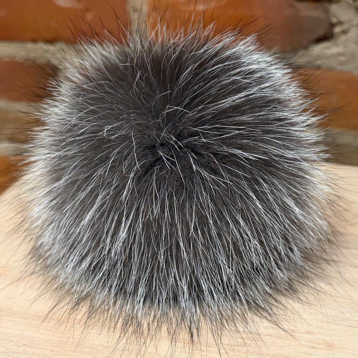 A round, fluffy silver fox-fur pom-pom in a mix of dark gray and silver-white tones, resting on a light wood surface with a blurred brick background.