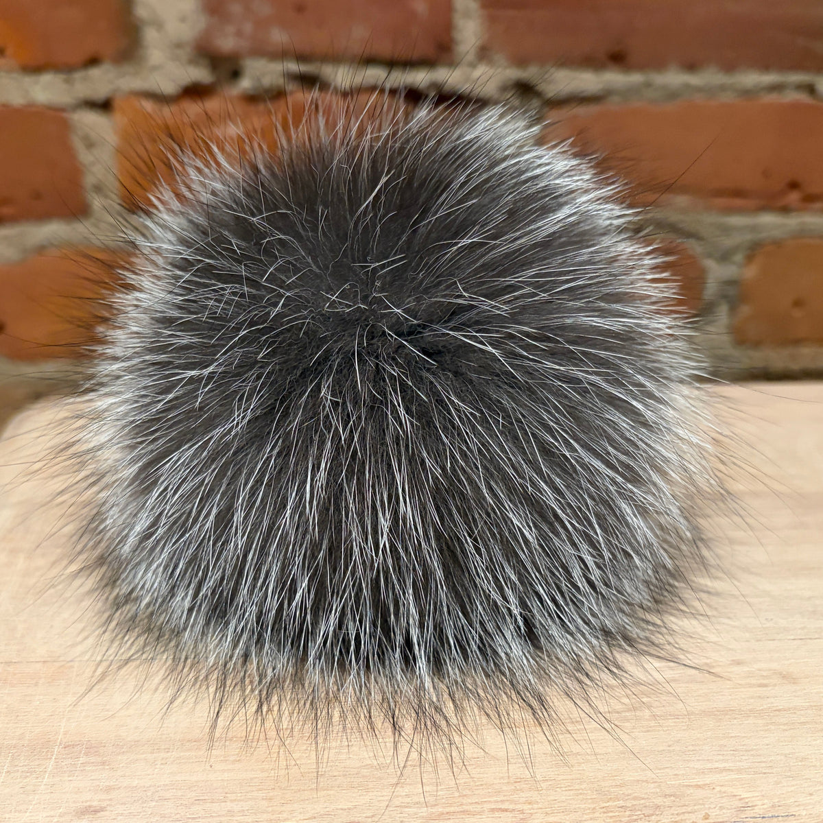 A round, fluffy silver fox-fur pom-pom in a mix of dark gray and silver-white tones, resting on a light wood surface with a brick wall background.