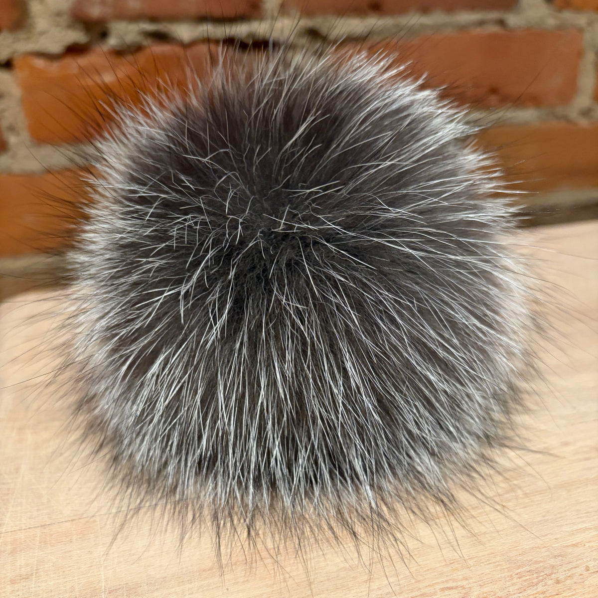A round, fluffy silver fox fur pom-pom in a mix of dark gray and silver-white fibers, resting on a light wood surface with a red brick wall in the background.