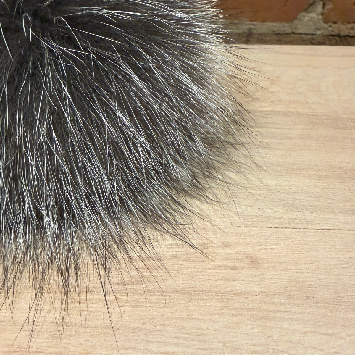 A close-up of a fluffy pom-pom made of dark gray and silver-toned silver fox fur, resting on a light wood surface with a brick wall in the background.
