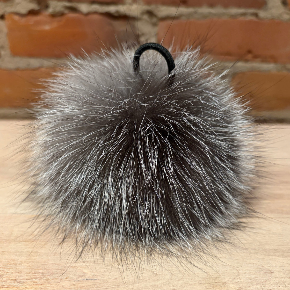 A fluffy gray and white silver fox fur pom-pom with a black elastic loop, resting on a light wood surface with a brick wall background.