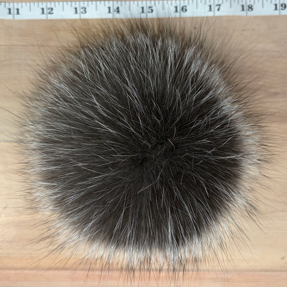 A round, fluffy silver fox fur pom-pom in dark brown and gray with lighter silver tips, shown on a light wood background with a measuring tape at the top for scale.