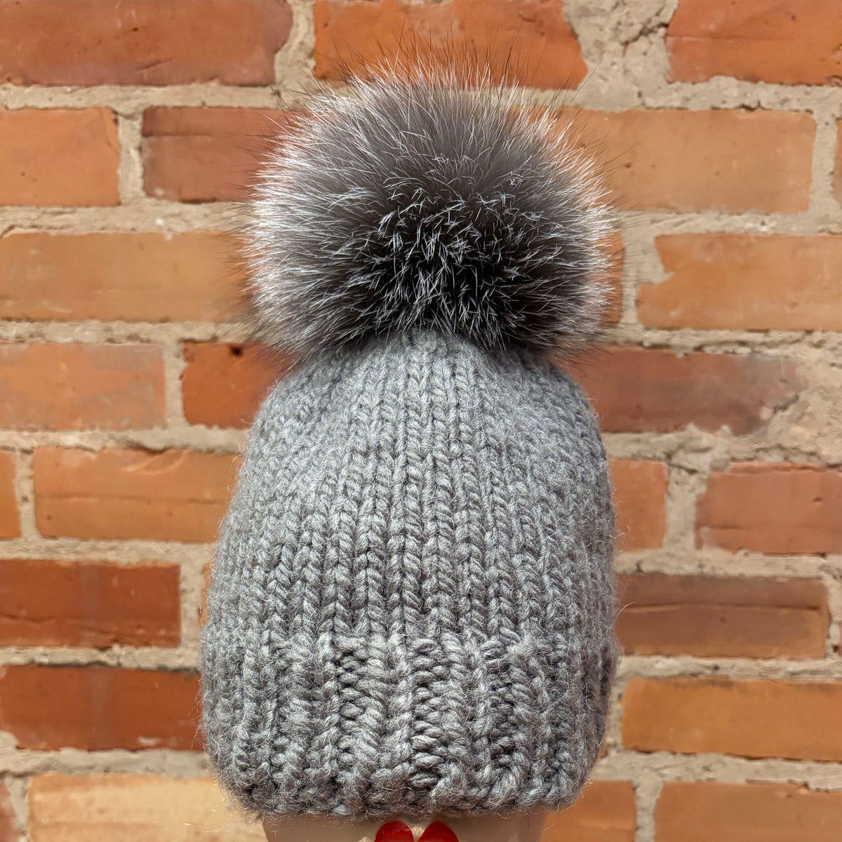 A gray chunky knit beanie with a large fluffy gray pom-pom, displayed on a mannequin head with red lipstick against a red brick wall background.