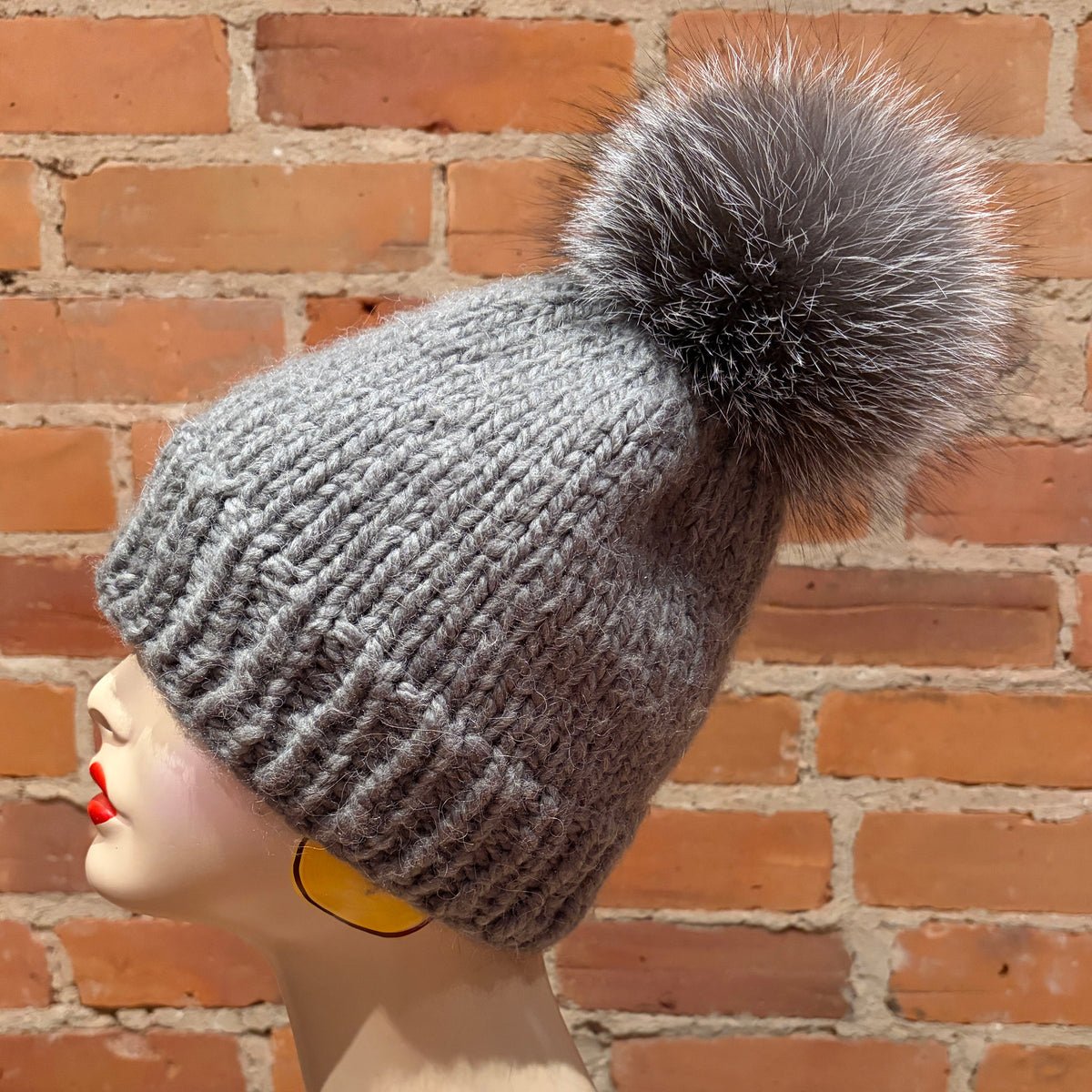 A gray ribbed knit beanie with a large fluffy pom-pom, displayed on a mannequin head in profile against a red brick wall background.