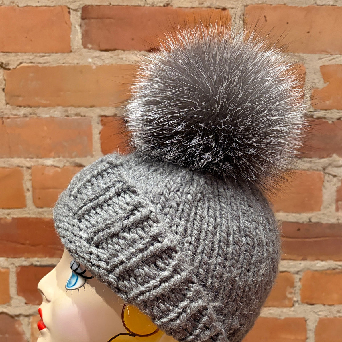 A gray knit beanie with a large fluffy gray-and-white pom-pom, displayed on a mannequin head against a red brick wall background.