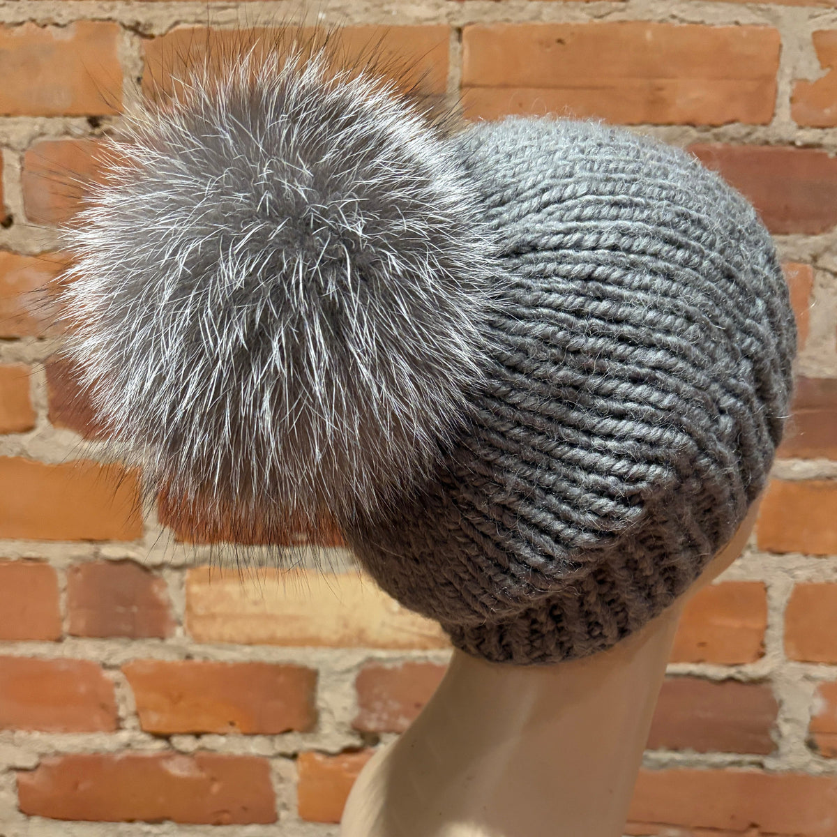 A gray ribbed knit beanie with a large fluffy silver-gray silver fox fur pom-pom, displayed on a beige mannequin head against a red brick wall background.