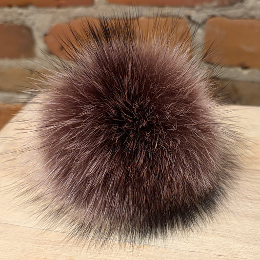 A round, fluffy pom-pom accessory made of dark brown and black raccoon fur or animal fur, resting on a light wood surface with a blurred brick wall background.