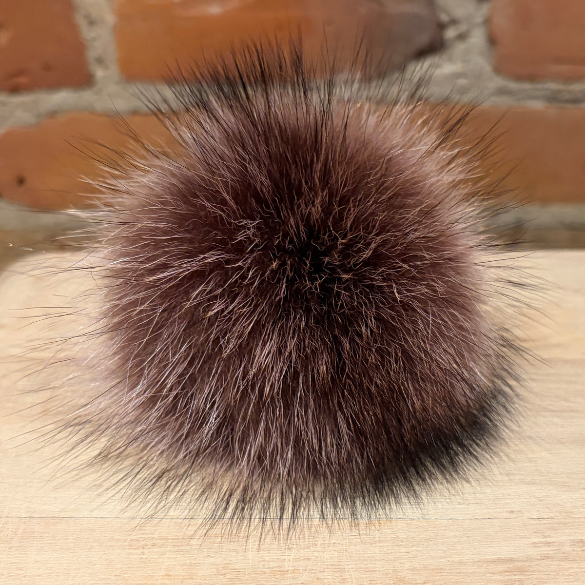 A round, fluffy raccoon-fur pom-pom in a dark brown and black mottled pattern, resting on a light wood surface with a blurred brick wall background.