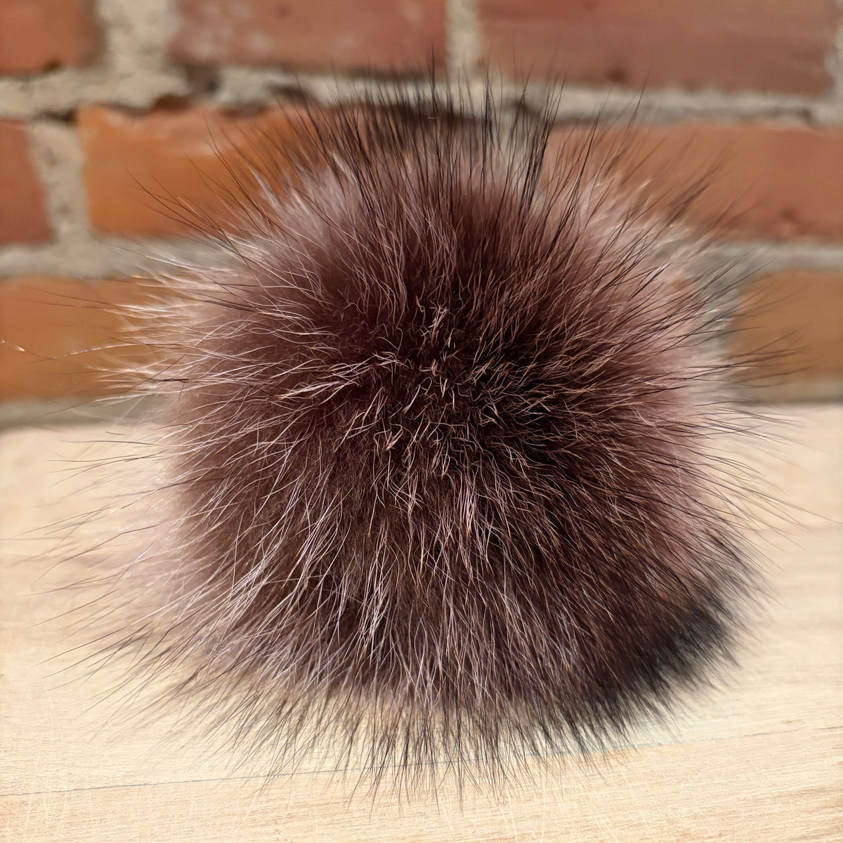 A round, fluffy raccoon fur pom-pom in a dark brown and black colorway, resting on a light wood surface with a blurred brick wall background.