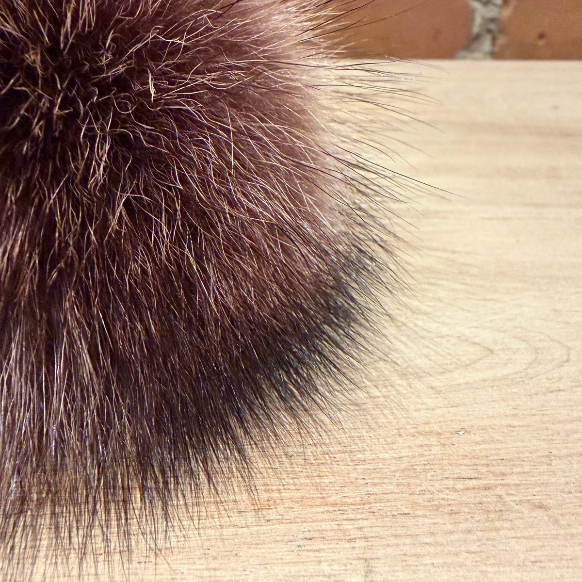 A close-up of a dark brown fluffy raccoon fur pom-pom resting on a light wood surface with a brick wall in the background.