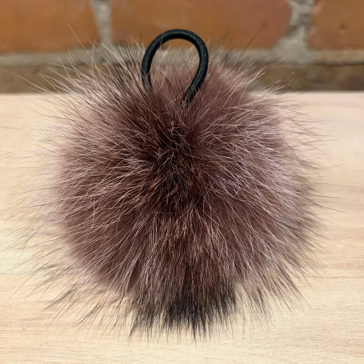 A fluffy raccoon-fur pom-pom keychain or bag charm in a dusty taupe-brown color, featuring a black elastic loop at the top, resting on a light wood surface with a brick wall in the background.