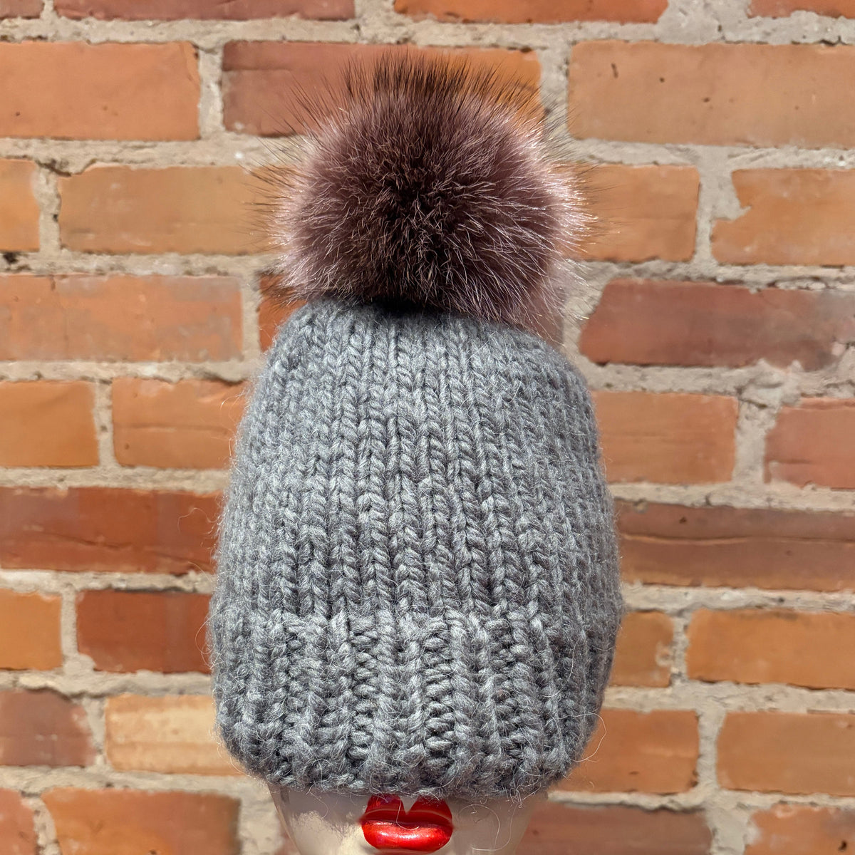 A gray ribbed knit beanie with a large brown raccoon fur pom-pom, displayed on a mannequin head with red lipstick in front of a brick wall background.