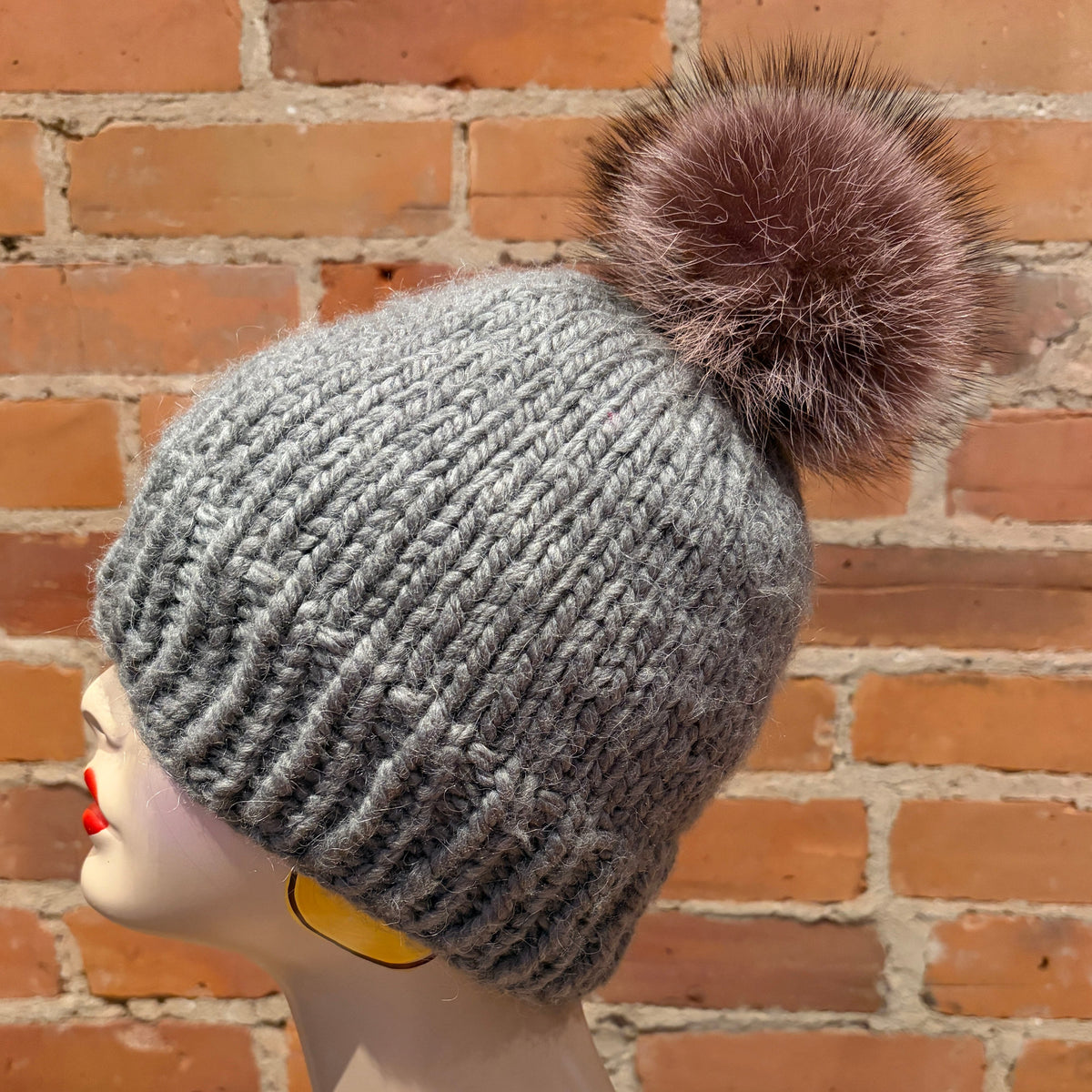 A gray ribbed knit beanie with a large brown raccoon-fur pom-pom, displayed on a mannequin head against a red brick wall background.