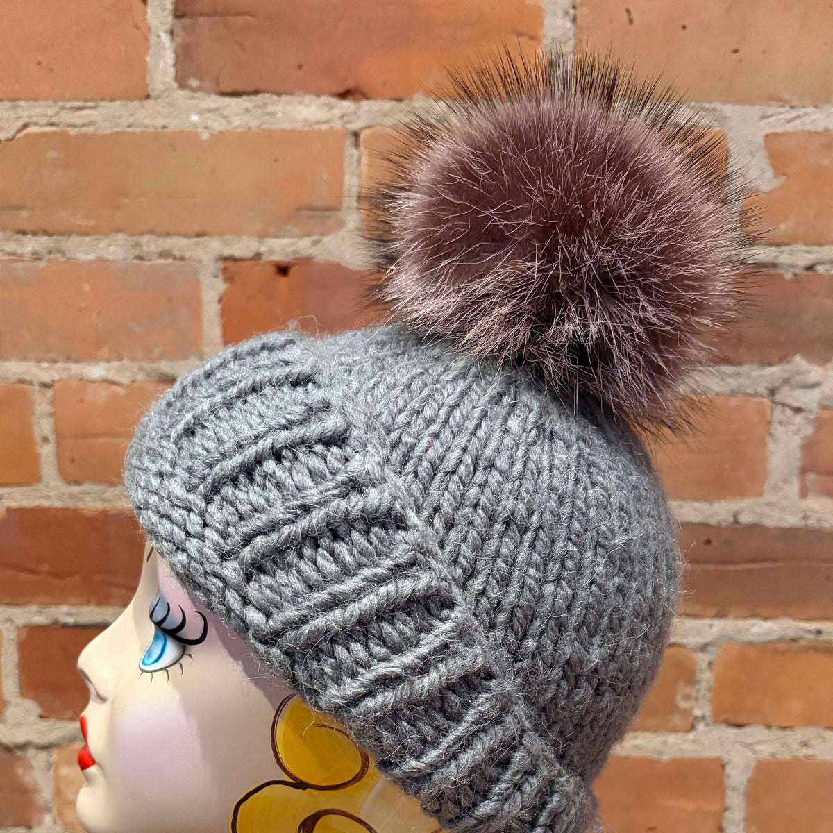 A gray ribbed knit beanie with a large brown raccoon-fur pom-pom, displayed on a mannequin head against a red brick wall background.