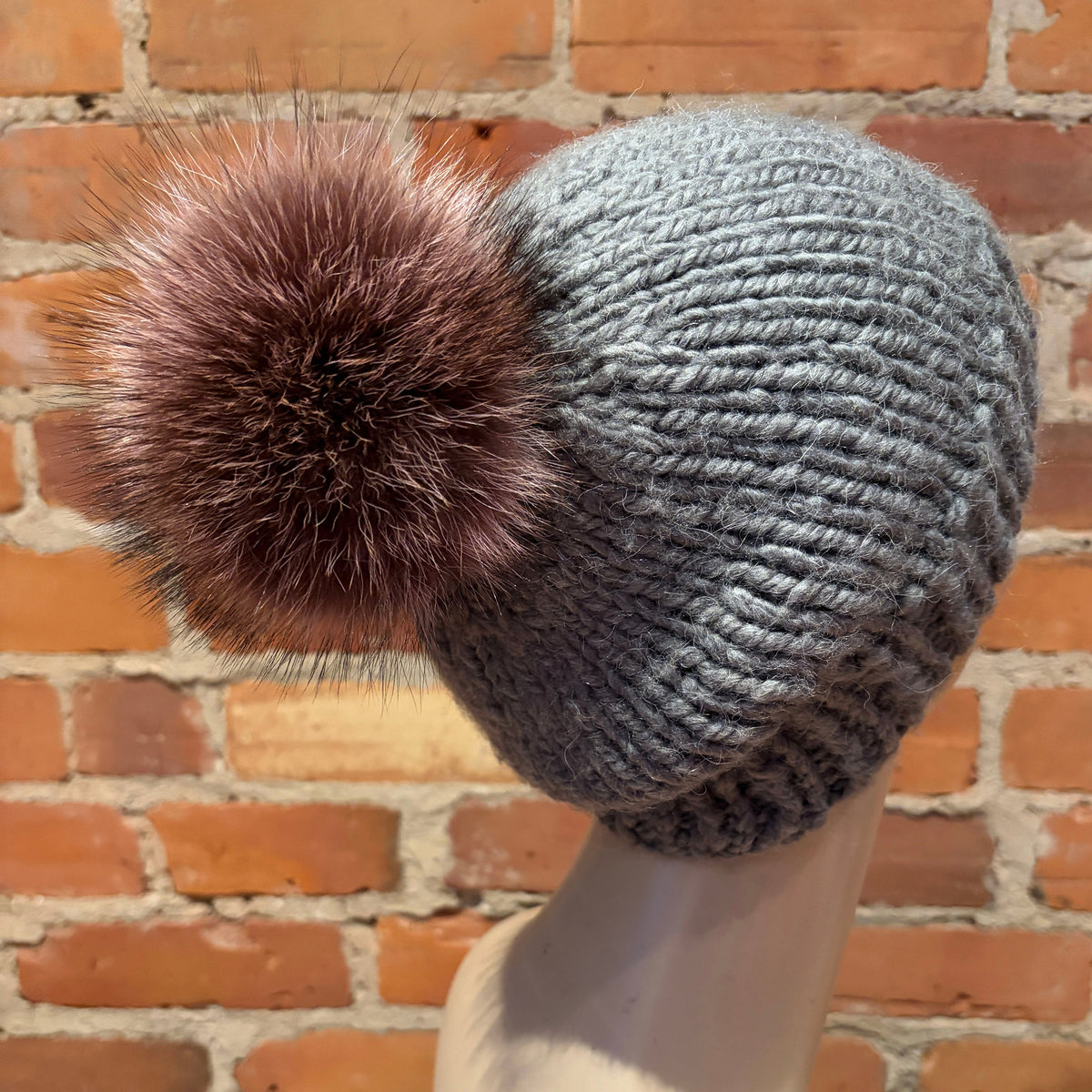 A gray ribbed knit beanie with a large brown raccoon fur pom-pom, displayed on a mannequin head against a red brick wall background.