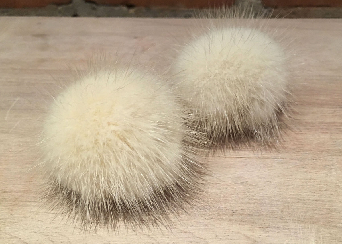 Two fluffy cream-colored mink fur pom poms placed on a light wood surface with a brick wall in the background.