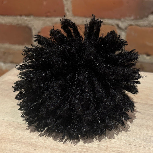 A black curly hat pom pomdisplayed on a light wood surface with a brick wall background.