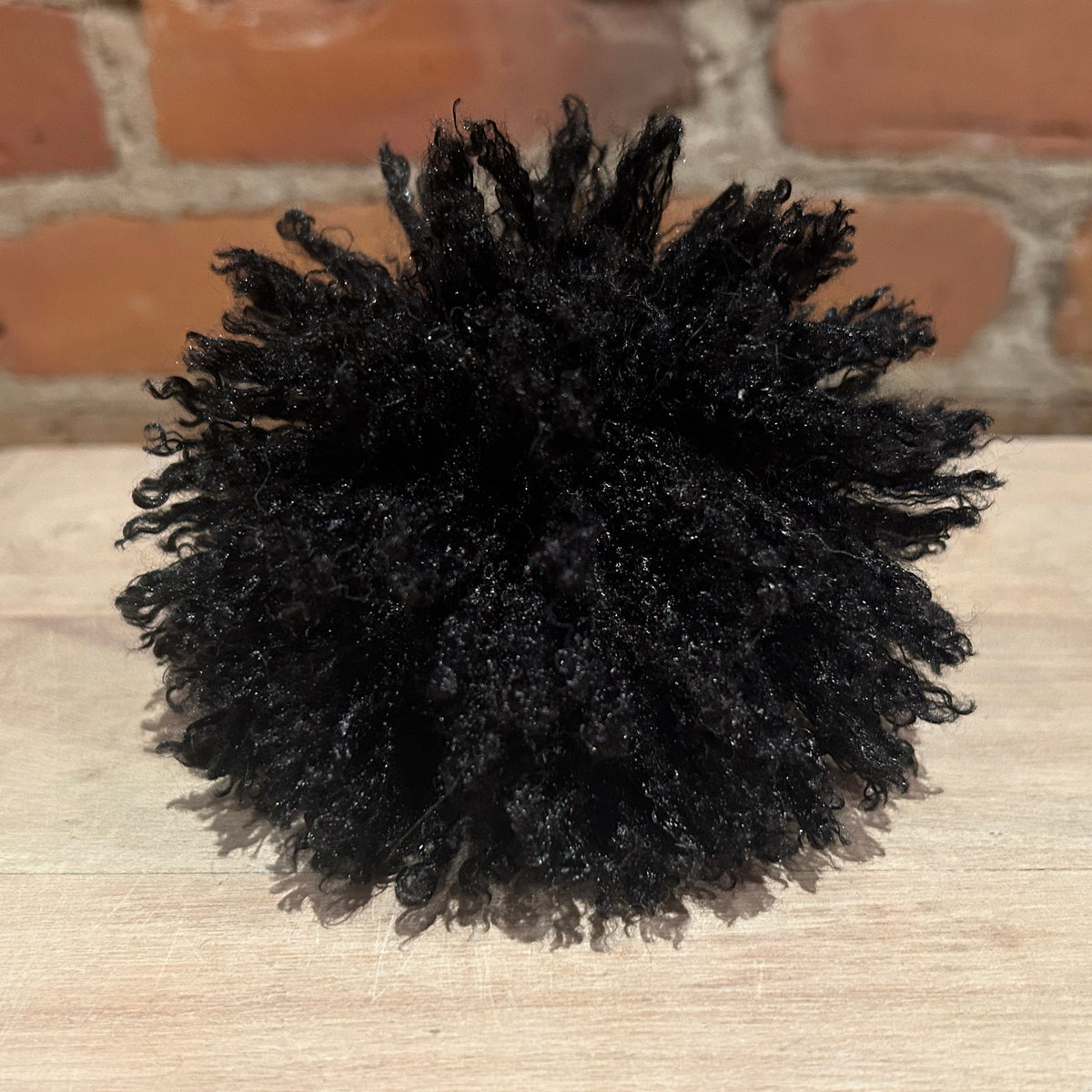 A black curly hat pom displayed on a light wood surface with a brick wall background.