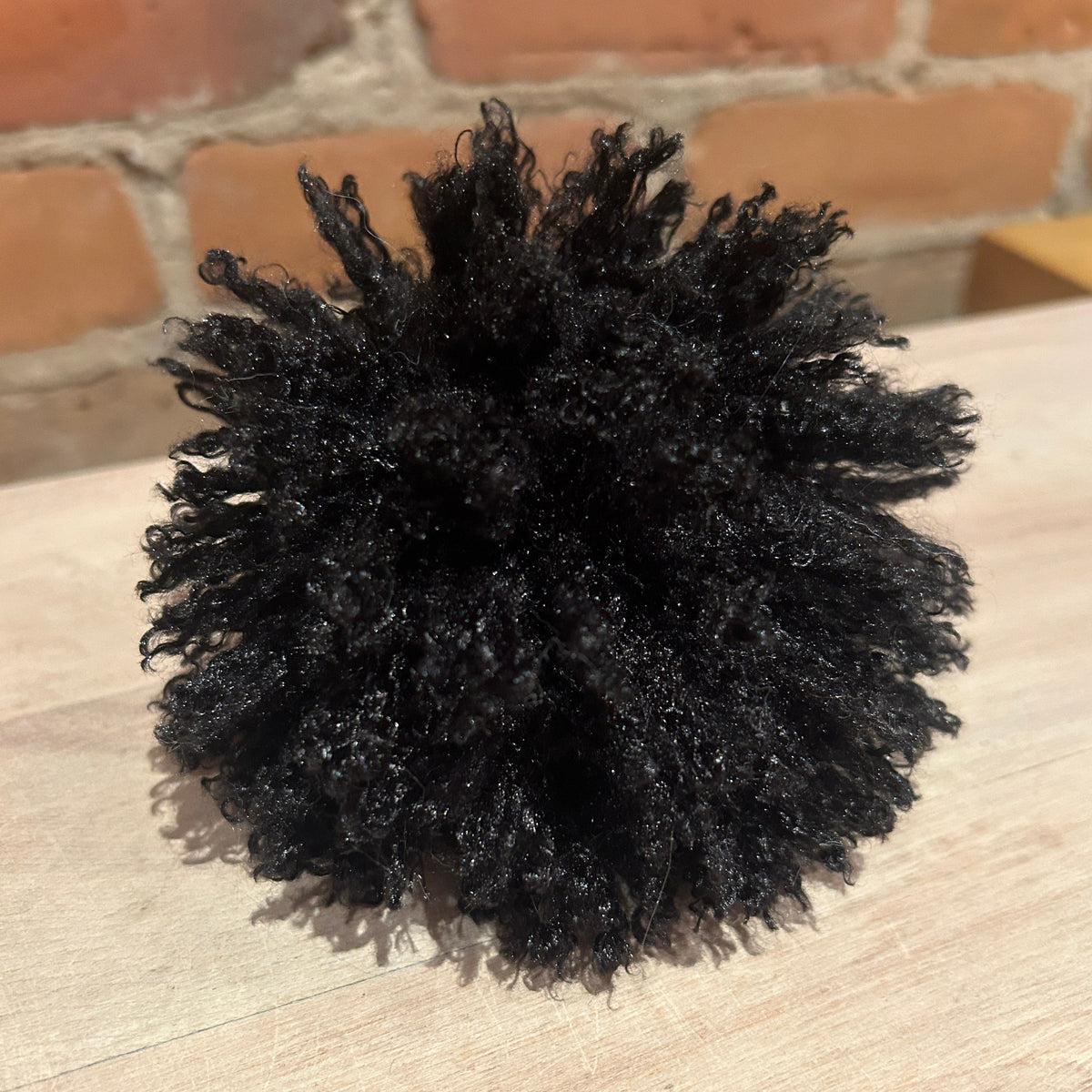 A black curly pom pom with a natural afro texture, displayed on a light wood surface with a blurred brick wall background.