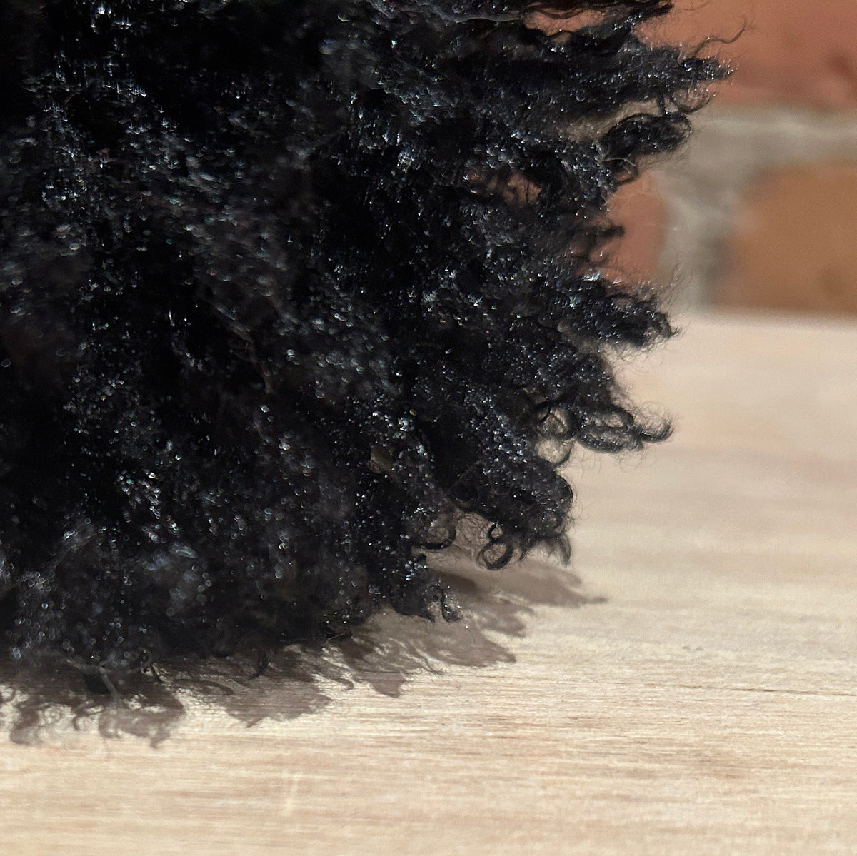 A close-up of a black curly hat pom resting on a light wood surface, with a blurred brick wall in the background.