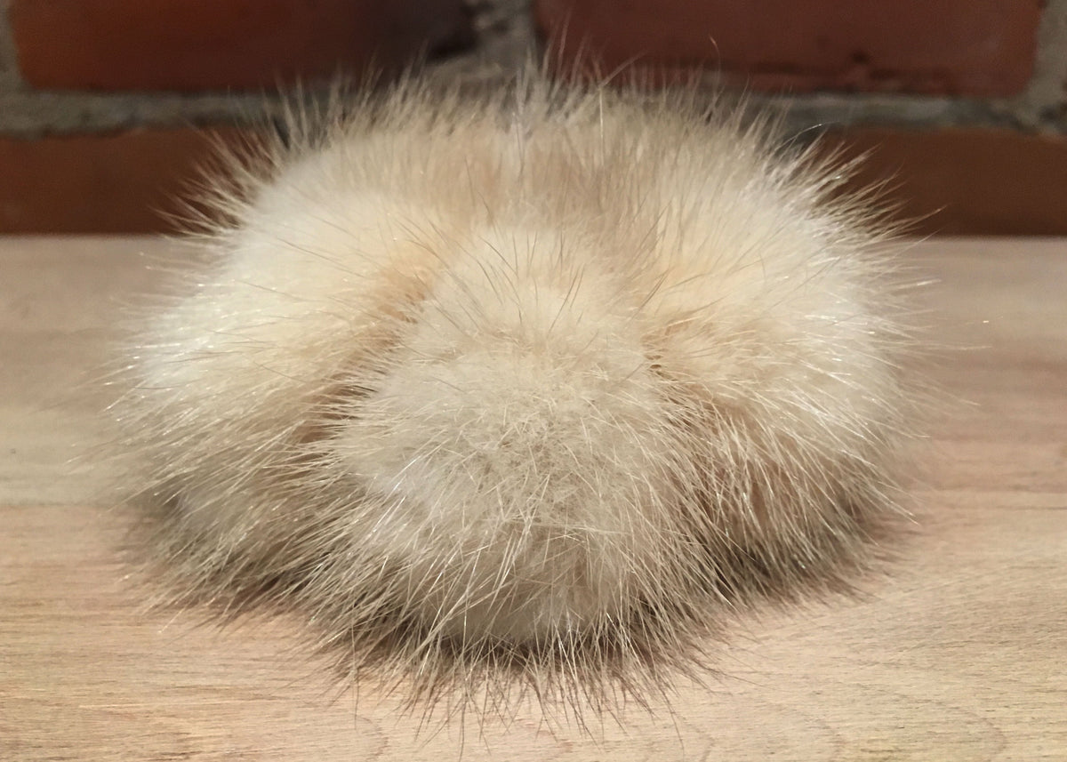 A fluffy mink fur pom-pom ball in a light beige color, resting on a light wood surface with a brick wall in the background.