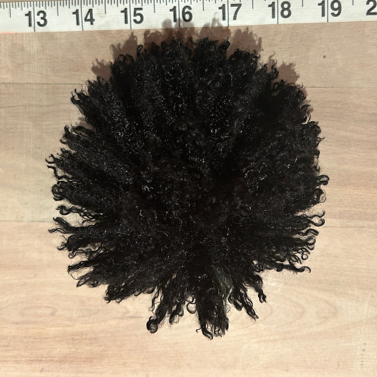 A black curly pom pom displayed on a light wood surface with a measuring tape ruler at the top to show scale.