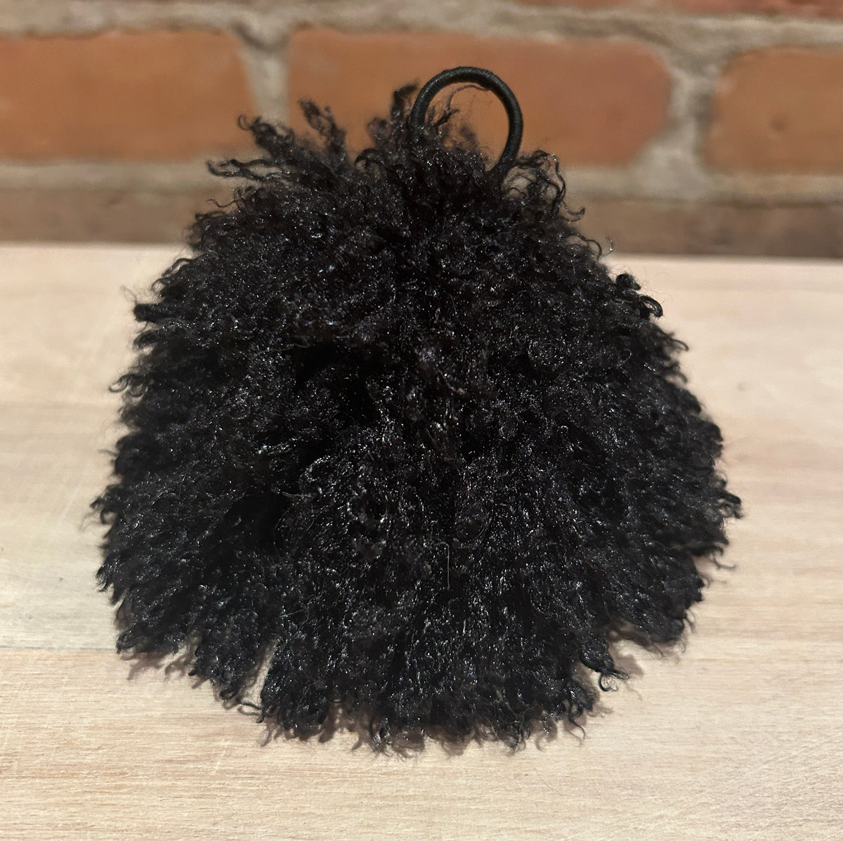 A black curly faux-fur pom-pom accessory with a black metal ring at the top, resting on a light wood surface with a brick wall in the background.
