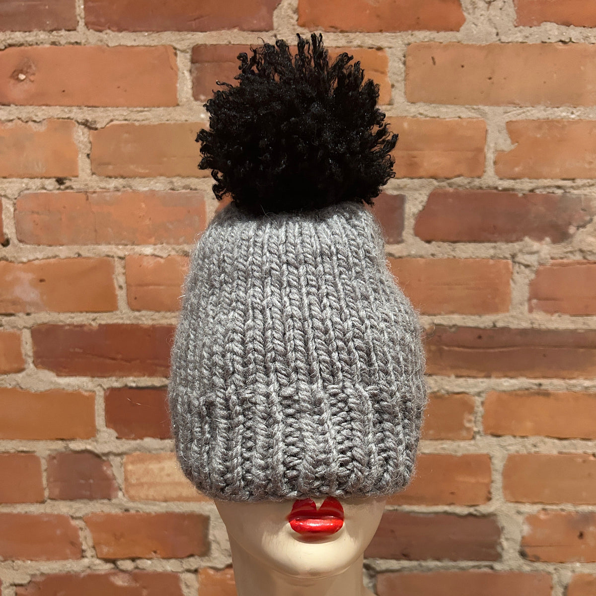 A light gray chunky knit beanie with a large black pom-pom, displayed on a mannequin head against a red brick wall background.