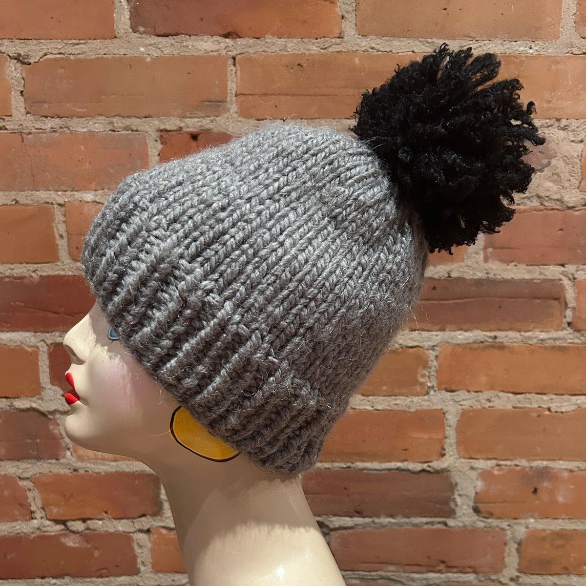 A gray ribbed knit beanie with a large black pom-pom, displayed on a mannequin head against a red brick wall background.