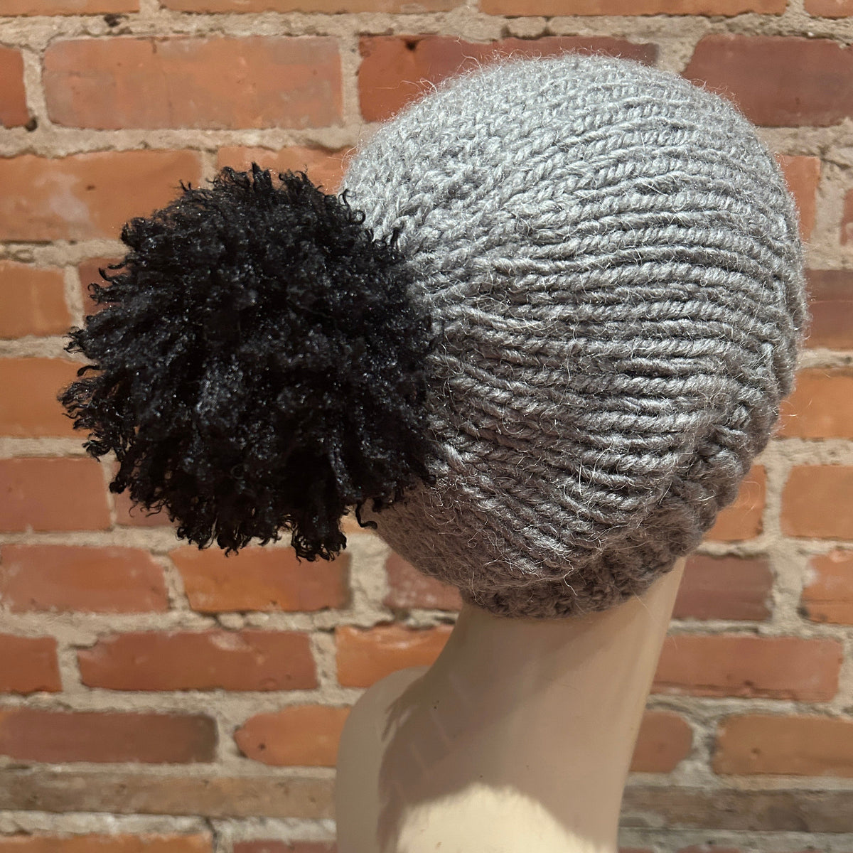 A gray ribbed knit beanie with a large black pom-pom, displayed on a mannequin head against a red brick wall background.