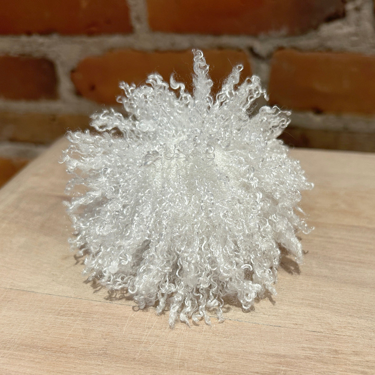 A white curly faux fur faux fur hat pom, photographed on a light wood surface with a brick wall background.