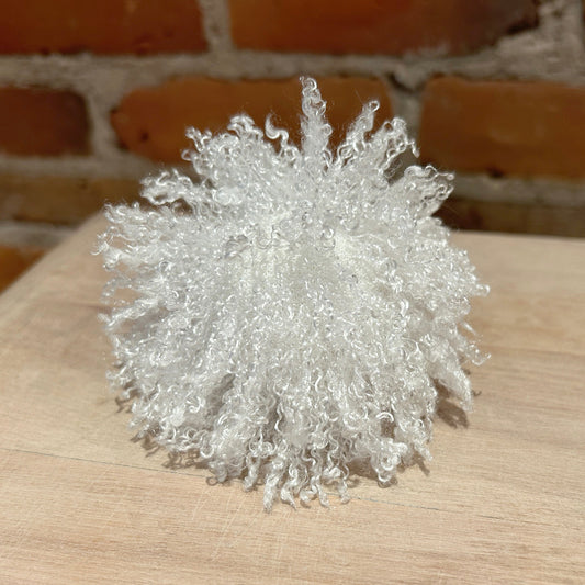 A white curly faux fur faux fur hat pom, photographed on a light wood surface with a brick wall background.