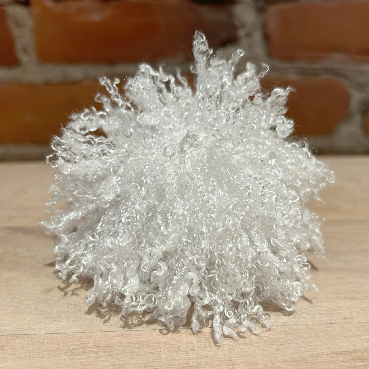 A fluffy, curly white decorative pom-pom or pom-pom cluster placed on a light wood surface with a blurred brick wall background.