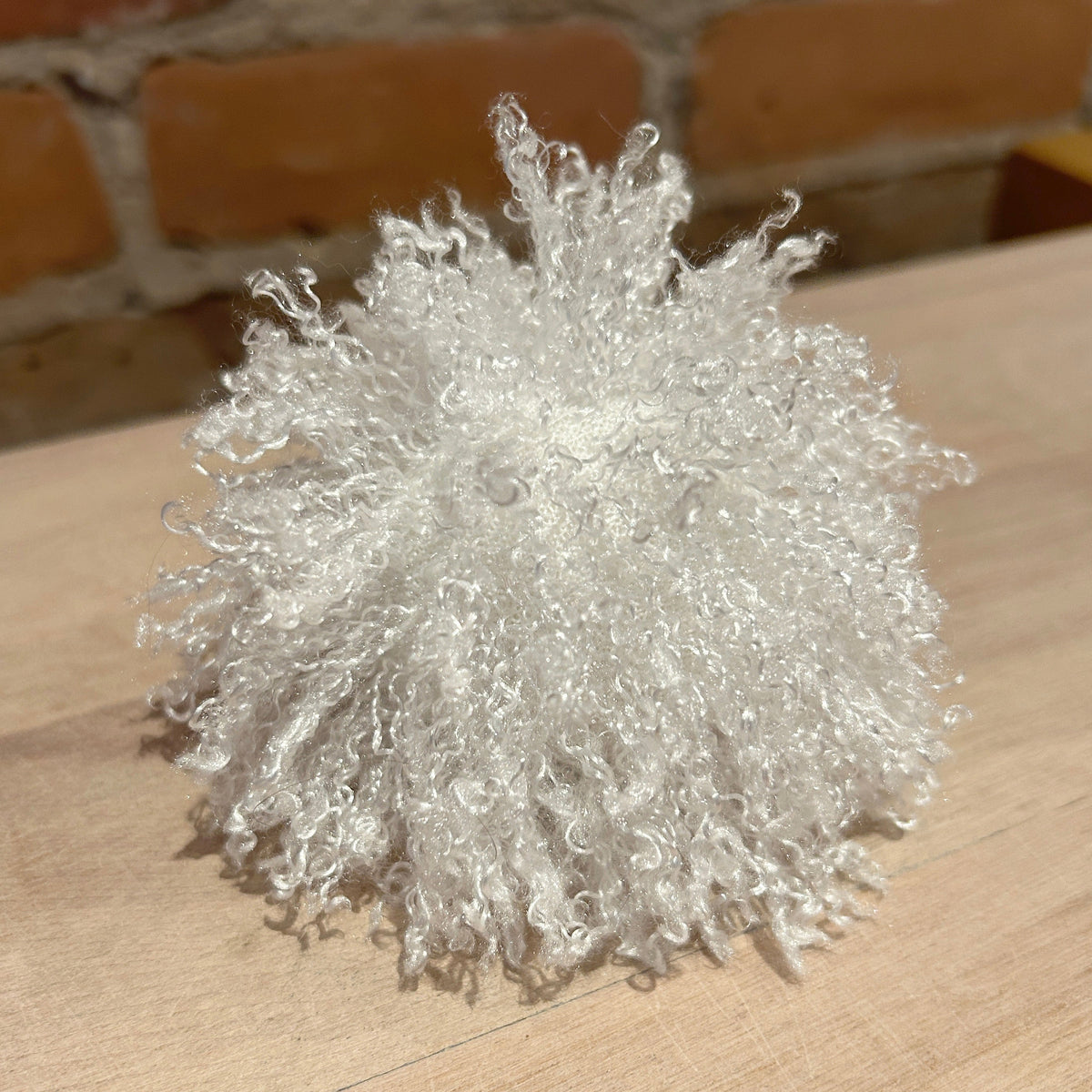 A fluffy, curly white decorative pom-pom or textured ball made of synthetic fibers, resting on a light wood surface with a blurred brick wall background.