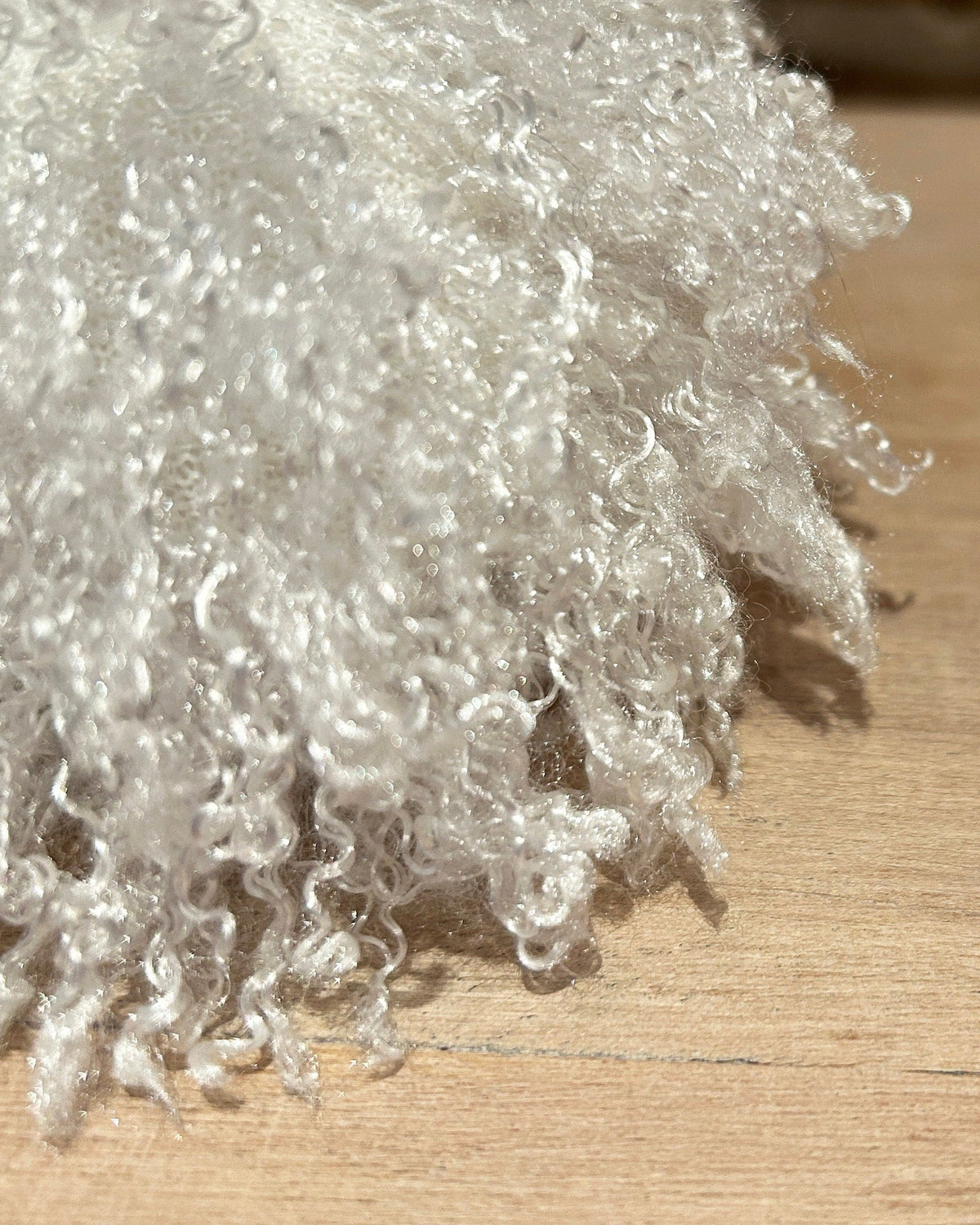 A close-up of a fluffy, curly white faux fur or sheepskin-style pom pom on a light wood surface with a blurred background.