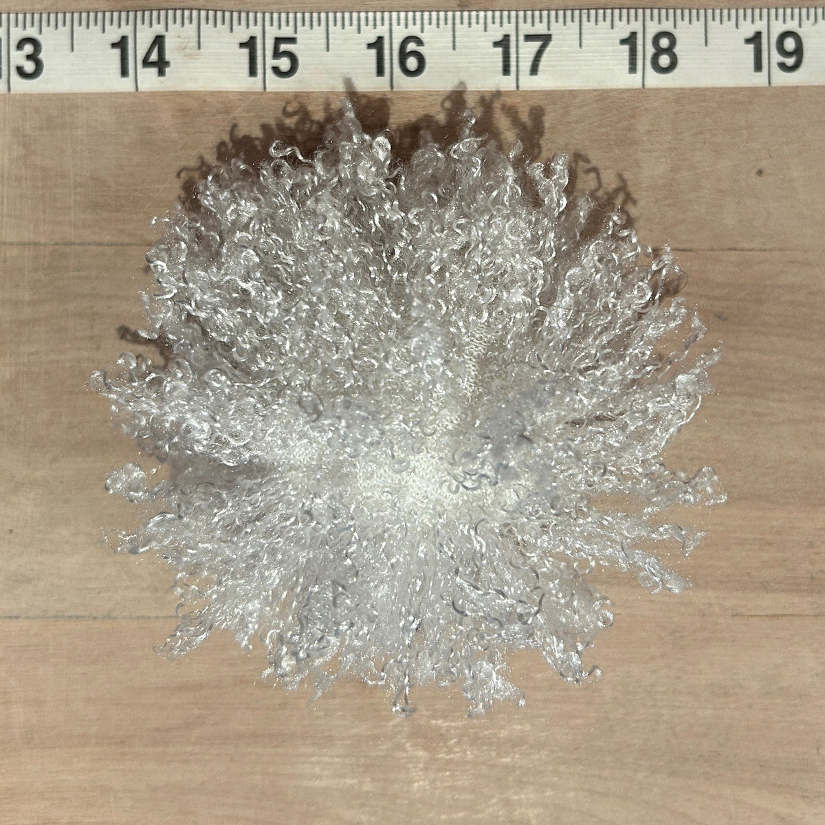 A white curly faux fur hat pom on a light wood surface with a measuring tape ruler visible at the top for scale.