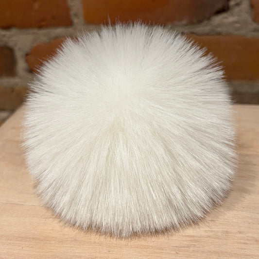 A fluffy white faux fur pom-pom ball resting on a light wood surface with a brick wall background.