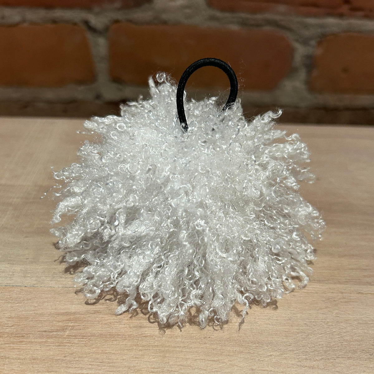 A white curly faux fur pom-pom ornament with a black hanging loop, resting on a light wood surface with a brick wall background.